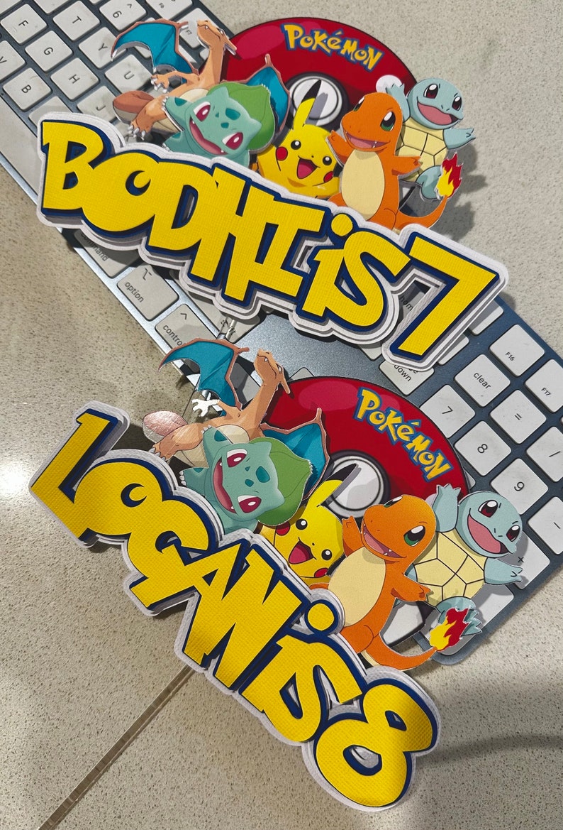 K&ouml;nnte beinhalten: Zwei mehrschichtige Tortenaufs&auml;tze mit Pok&eacute;mon-Charakteren und den Texten "BODHI IS 7" und "LOGAN IS 8". Die Aufs&auml;tze zeigen Pikachu, Glumanda, Bisasam, Schiggy und Glurak, mit einem roten Pok&eacute;mon-Logo.