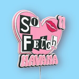 Può includere: Una decorazione a forma di cuore rosa a strati con le parole "So Fetch" in bianco e nero, un'impronta di labbra rosse e la parola "Havana" in rosa. La decorazione è montata su un sottile filo d'argento.