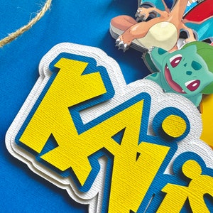 K&ouml;nnte beinhalten: Ein mehrschichtiger Papier-Kuchenaufsatz mit dem Namen "Kai" in Gelb und Blau, mit Zeichentrickfiguren. Der Aufsatz befindet sich auf einem blauen Hintergrund mit einer Schnur aus Bindfaden. Zu den Figuren geh&ouml;rt ein drachenartiges Wesen mit orangefarbenen und blauen Fl&uuml;geln.