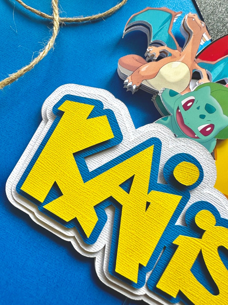 K&ouml;nnte beinhalten: Ein mehrschichtiger Papier-Kuchenaufsatz mit dem Namen "Kai" in Gelb und Blau, mit Zeichentrickfiguren. Der Aufsatz befindet sich auf einem blauen Hintergrund mit einer Schnur aus Bindfaden. Zu den Figuren geh&ouml;rt ein drachenartiges Wesen mit orangefarbenen und blauen Fl&uuml;geln.