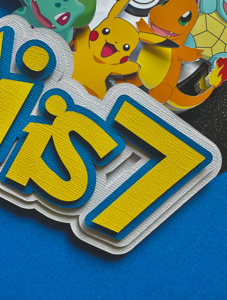 K&ouml;nnte beinhalten: Mehrschichtige Papierdekoration mit dem Text "Happy 7" in Gelb und Blau, vor blauem Hintergrund. Die Dekoration zeigt Bilder von Pikachu, Glumanda und anderen Pok&eacute;mon-Charakteren.