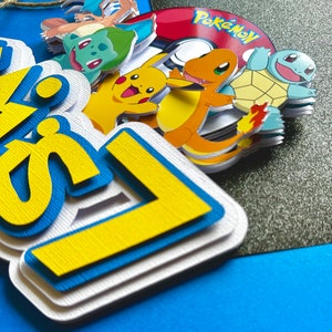 K&ouml;nnte beinhalten: Ein mehrschichtiger Tortenaufsatz aus Papier mit dem Wort "Happy" in Gelb und Blau und einem wei&szlig;en Rand. Der Aufsatz enth&auml;lt mehrere Pok&eacute;mon-Charaktere und einen rot-wei&szlig;en Pok&eacute;ball.
