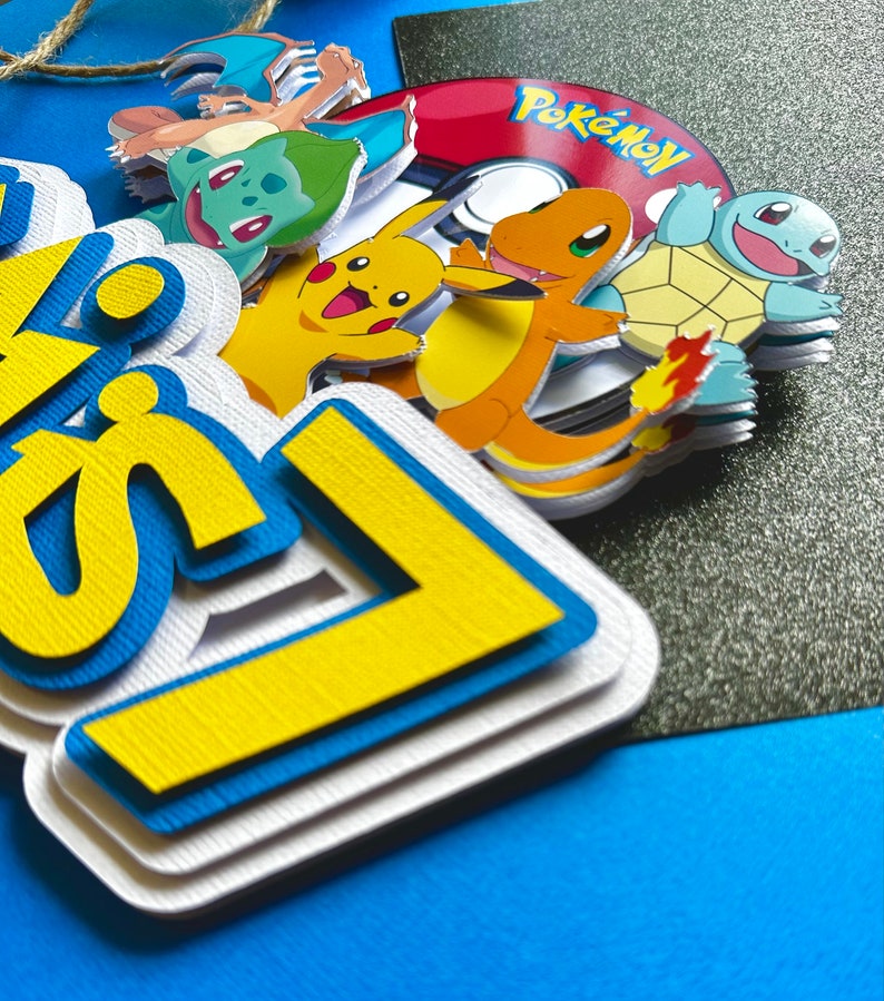 K&ouml;nnte beinhalten: Ein mehrschichtiger Tortenaufsatz aus Papier mit dem Wort "Happy" in Gelb und Blau und einem wei&szlig;en Rand. Der Aufsatz enth&auml;lt mehrere Pok&eacute;mon-Charaktere und einen rot-wei&szlig;en Pok&eacute;ball.
