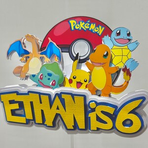 K&ouml;nnte beinhalten: Bunte Pok&eacute;mon-Tortenaufsatz mit Charizard, Bulbasaur, Pikachu, Charmander und Squirtle. Das Design umfasst einen rot-wei&szlig;en Pok&eacute;ball und den Text "Ethan is 6" in Gelb und Blau.