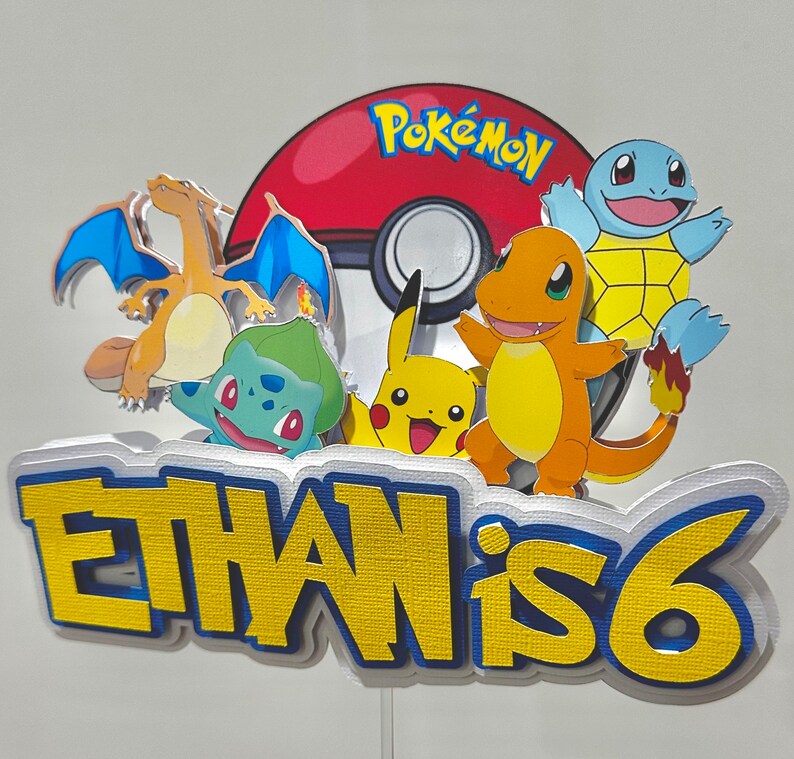 K&ouml;nnte beinhalten: Bunte Pok&eacute;mon-Tortenaufsatz mit Charizard, Bulbasaur, Pikachu, Charmander und Squirtle. Das Design umfasst einen rot-wei&szlig;en Pok&eacute;ball und den Text "Ethan is 6" in Gelb und Blau.