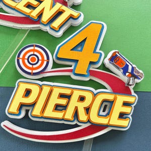 May include: A layered cake topper with the name "Pierce" in yellow letters with a red and white accent. The number "4" is in orange and blue, with a target and toy gun. The background is green and blue.