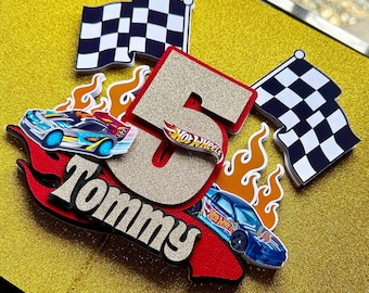 May include: A birthday cake topper featuring a gold glitter number 5 with a Hot Wheels logo, a red and black base with the name "Tommy", and two race cars. Includes checkered flags and flame accents.