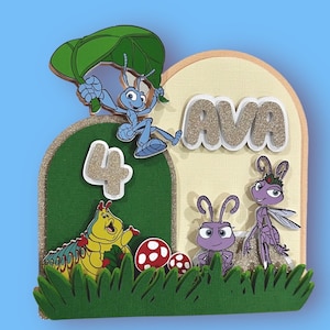 Può includere: Topper per torta di compleanno colorato con personaggi del film d'animazione "A Bug's Life". Il design include il nome "AVA" in lettere glitterate, il numero "4" e vari personaggi di insetti. Lo sfondo è verde e beige.