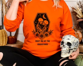 May include: Orange sweatshirt with a black graphic of two skeletons embracing inside a coffin with sunflowers. The text "Meet me at the graveyard" is printed below the graphic.