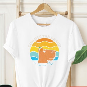 May include: White t-shirt with a retro-style graphic of a capybara wearing a slice of orange and the text "Commit Tax Fraud".