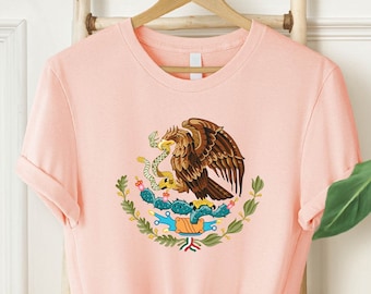 May include: A pink t-shirt with a graphic of the Mexican coat of arms featuring a golden eagle perched on a cactus with a snake in its beak. The eagle is surrounded by green leaves and a banner with the words "Estados Unidos Mexicanos".