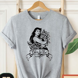 May include: A gray t-shirt with a black and white illustration of a woman holding flowers. The text "Como La Flor" is printed on the shirt.