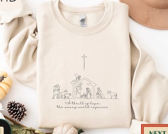 May include: Sand-colored sweatshirt featuring a minimalist line drawing of a nativity scene with a star above. The text "A thrill of hope, the weary world rejoices" is below the drawing. The sweatshirt is a neutral color.