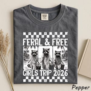 May include: Pepper-colored t-shirt with a graphic featuring three raccoons wearing sunglasses and carrying luggage, with the text "FERAL & FREE GIRLS TRIP 2026". The shirt has a crew neck and a Comfort Colors tag.