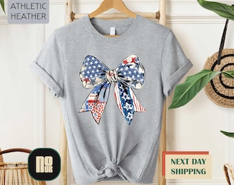 May include: Athletic heather gray t-shirt featuring a patriotic bow design. The bow incorporates red, white, and blue elements with stars and stripes. The shirt is a casual wear item.