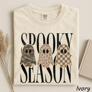 May include: An ivory t-shirt featuring the black text SPOOKY SEASON and three ghost illustrations. The ghosts have distinct patterns: one with green and brown camouflage, one with a tan leopard print, and one with a brown and tan checkerboard pattern.