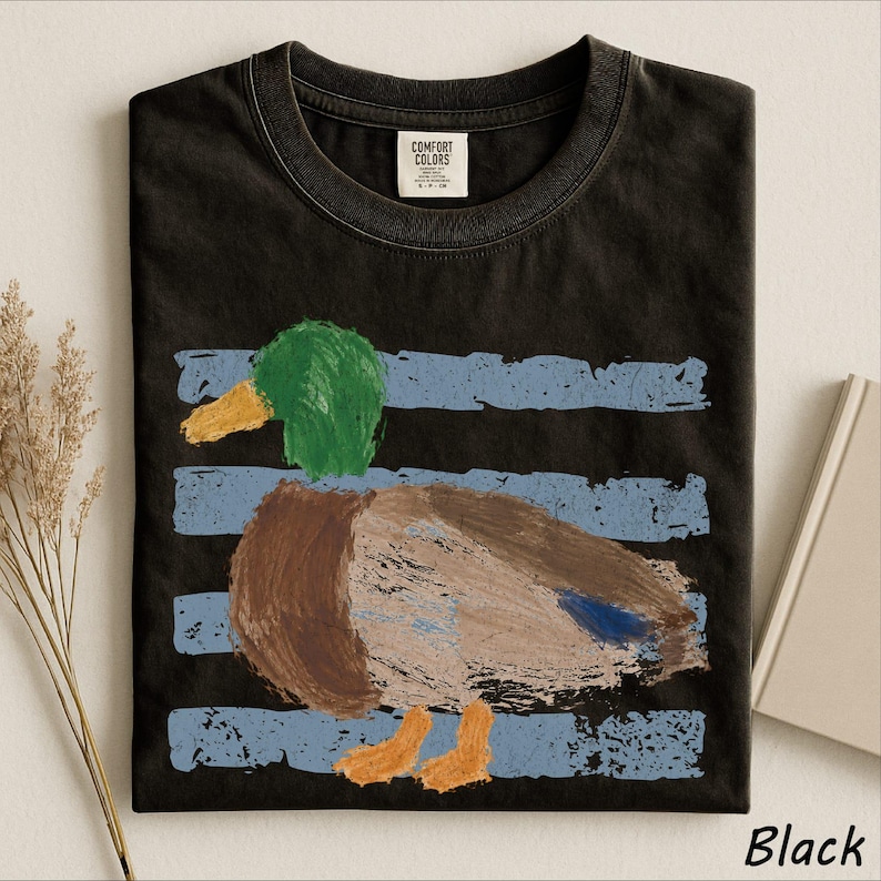 A black t-shirt with a colorful painting of a duck on it, along with some dried flowers and a book.