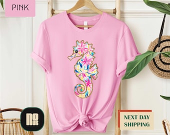 May include: Pink t-shirt featuring a colorful seahorse design. The seahorse is decorated with floral patterns in shades of pink, blue, yellow, and gold. The shirt has a round neck and short sleeves. The word "PINK" is in a pink box.
