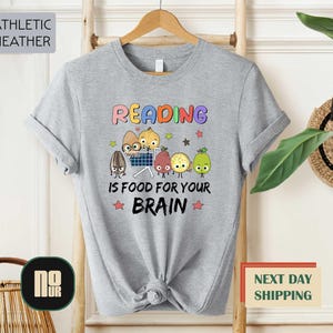 May include: Athletic heather gray t-shirt with the text "Reading is Food for Your Brain" in colorful letters. The graphic features cartoon fruits and vegetables with faces, including a sunflower seed, onion, pear, and plum.