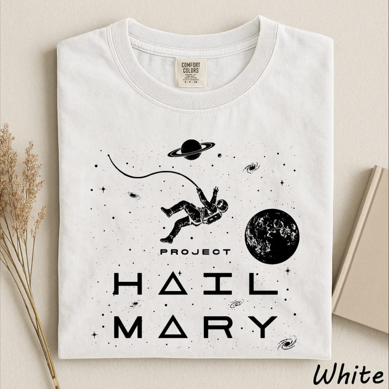 May include: A white short-sleeved t-shirt featuring a black graphic print of an astronaut floating in space with a tether, a planet with rings, and a moon. The text "PROJECT HAIL MARY" is printed in black below the astronaut in a stylized font.