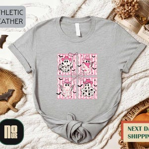 a gray t-shirt with a graphic design of four cartoon bats wearing hats and holding bats, arranged in a square pattern. The t-shirt is displayed on a white background, surrounded by various Halloween-themed items such as bats, a pumpkin, and a woven basket.