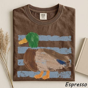 a brown t-shirt with a colorful drawing of a duck on it, along with some dried flowers and a book.