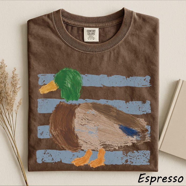 a brown t-shirt with a colorful drawing of a duck on it, along with some dried flowers and a book.