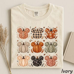 May include: An ivory t-shirt featuring a grid of nine pumpkin illustrations, each with mouse-ear shapes. The pumpkins display various patterns including leopard print, plaid, floral, polka dots, and solid colors. The neck label reads Comfort Colors.