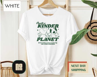 May include: White t-shirt with a green graphic that reads "Create a Kinder Planet." The design features a heart and a globe holding hands. The text "The world could always use more kindness" is below the graphic.