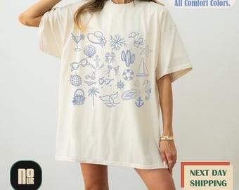 May include: Cream-colored t-shirt with a blue summer-themed graphic. The design includes illustrations of a cocktail, sunglasses, a seashell, a crab, a cactus, a sailboat, and other beach-related icons. Text on the shirt reads "All Sizes. All Colors. All Comfort Colors."