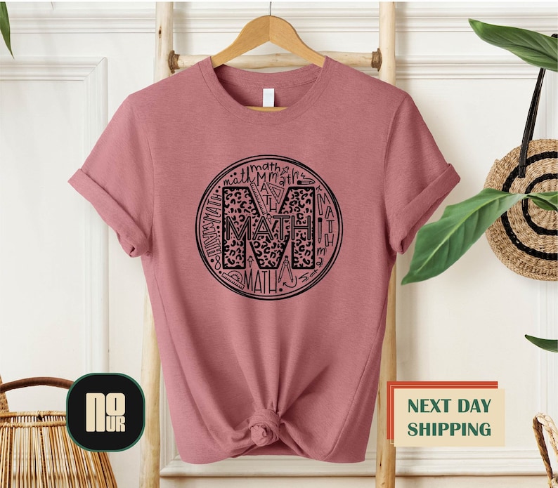 Puede incluir: Una camiseta rosa con un dise&ntilde;o circular negro que presenta la letra "M" rodeada de la palabra "MATH" en un patr&oacute;n de estampado de leopardo.