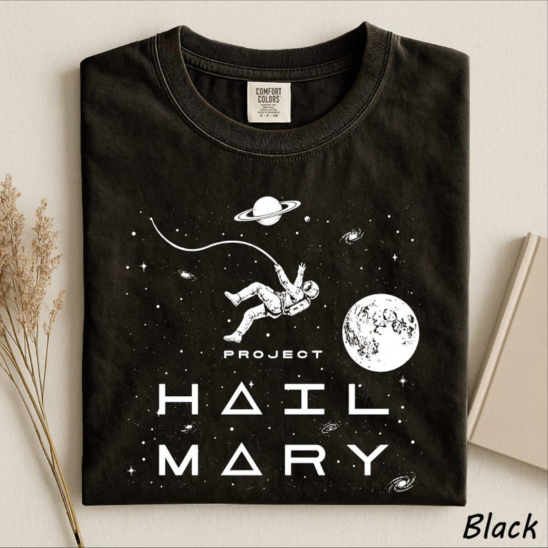 May include: A black t-shirt featuring a white graphic of an astronaut floating in space, tethered to a planet with rings. The design includes a large moon, stars, galaxies, and the text "PROJECT HAIL MARY" printed in a bold, modern white font.