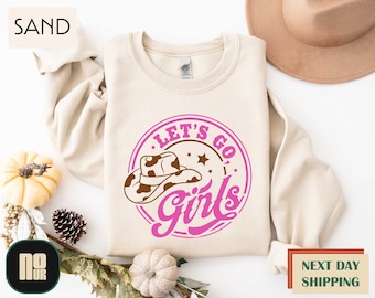 May include: Sand-colored sweatshirt with a circular graphic that reads "LET'S GO Girls" in pink and brown. The graphic includes a cowboy hat and stars. The sweatshirt is displayed on a neutral background with fall-themed decorations.