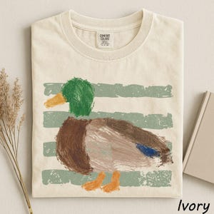 A white t-shirt with a colorful painting of a duck on it, along with some dried flowers and a book.
