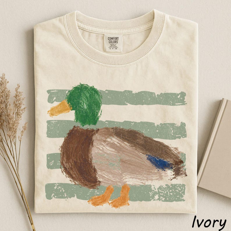 A white t-shirt with a colorful painting of a duck on it, along with some dried flowers and a book.