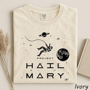 May include: An ivory t-shirt featuring a black graphic of an astronaut floating in space with a tether, a planet with rings, a moon, and stars. The text reads PROJECT HAIL MARY in a stylized font. The shirt has a Comfort Colors brand neck label.