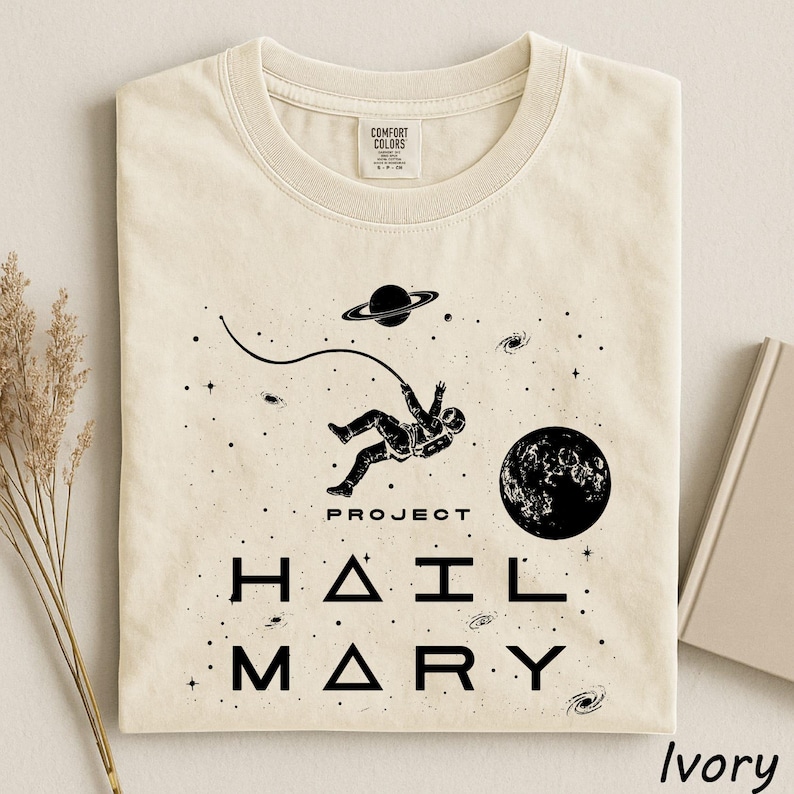 May include: An ivory t-shirt featuring a black graphic of an astronaut floating in space with a tether, a planet with rings, a moon, and stars. The text reads PROJECT HAIL MARY in a stylized font. The shirt has a Comfort Colors brand neck label.