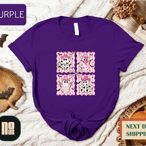 a purple t-shirt with a graphic design of four cartoon-style ghost characters, each wearing a pink hat and bow tie. The t-shirt is displayed on a white background, surrounded by various Halloween-themed items such as bats, pumpkins, and a book.