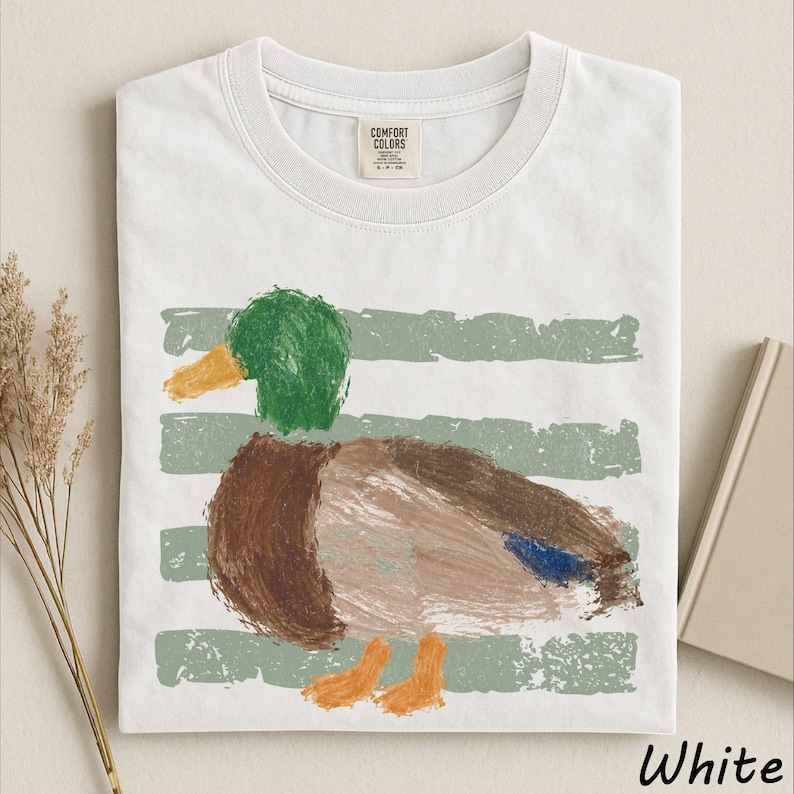 A white t-shirt with a colorful painting of a duck on it, along with some dried flowers and a book.