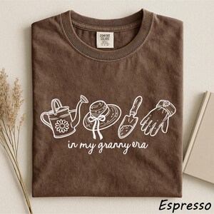 A brown t-shirt with a graphic design featuring a watering can, a hat, and gardening tools, along with the text "in my granny era" printed on it.