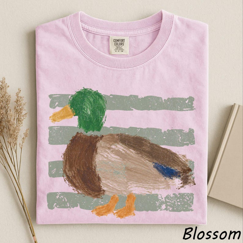 A pink t-shirt with a colorful painting of a duck on it, along with some dried flowers and a book.