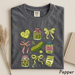 a gray t-shirt with a variety of colorful illustrations, including jars of pickles, a heart, a bow, a jar of chips, and a bag of candy.