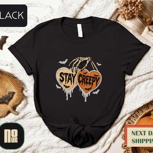 a black t-shirt with a Halloween-themed graphic design, including a skeleton hand holding a heart with the text "STAY CREEPY" and bats. The t-shirt is displayed on a white background, surrounded by Halloween-themed items such as bats, pumpkins, and a book.
