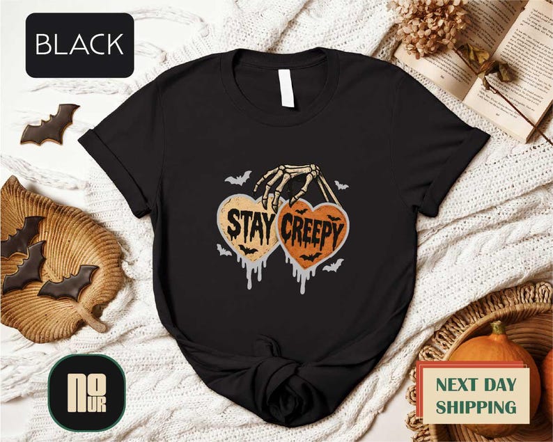 a black t-shirt with a Halloween-themed graphic design, including a skeleton hand holding a heart with the text "STAY CREEPY" and bats. The t-shirt is displayed on a white background, surrounded by Halloween-themed items such as bats, pumpkins, and a book.