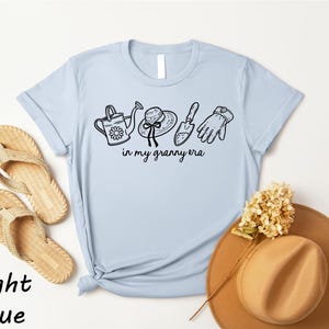 A light blue t-shirt with a graphic design of gardening tools and accessories, along with a pair of flip-flops and a hat, all placed on a white background.