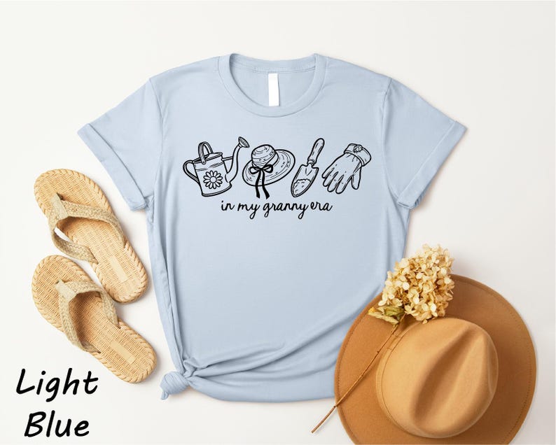 A light blue t-shirt with a graphic design of gardening tools and accessories, along with a pair of flip-flops and a hat, all placed on a white background.