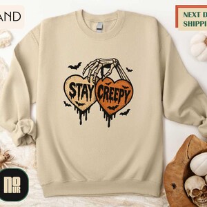 a beige sweatshirt with a Halloween-themed graphic design, including a skeleton hand holding a heart with the text "STAY CREEPY" and bats. The sweatshirt is displayed on a white background, and there are other Halloween-related items such as a pumpkin and a skull.