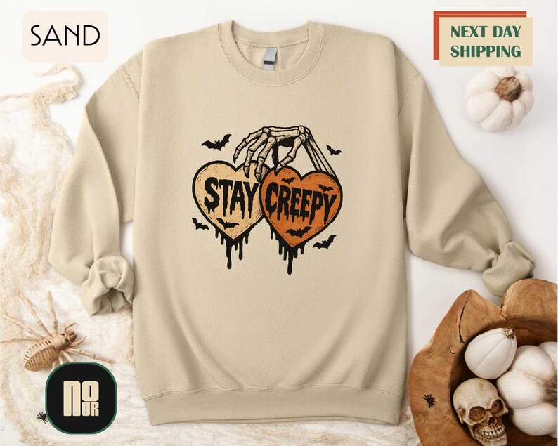 a beige sweatshirt with a Halloween-themed graphic design, including a skeleton hand holding a heart with the text "STAY CREEPY" and bats. The sweatshirt is displayed on a white background, and there are other Halloween-related items such as a pumpkin and a skull.