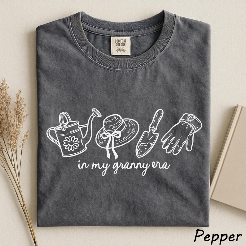 A gray t-shirt with a graphic design printed on it, featuring a watering can, a hat, a shovel, and a pair of gloves, along with the text "in my granny era".