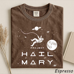 May include: A brown t-shirt featuring a white graphic of an astronaut floating in space with a planet, a moon, and stars. The text "PROJECT HAIL MARY" is printed in white below the astronaut. The shirt has a crew neckline and a Comfort Colors label.